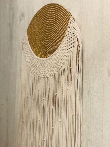 Decoración de pared en macramé con un diseño circular y flecos en tonos claros.
